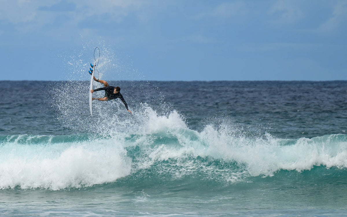 Surfer performing aerial trick on wave with FCS II compatible surfboard fins, ocean backdrop
